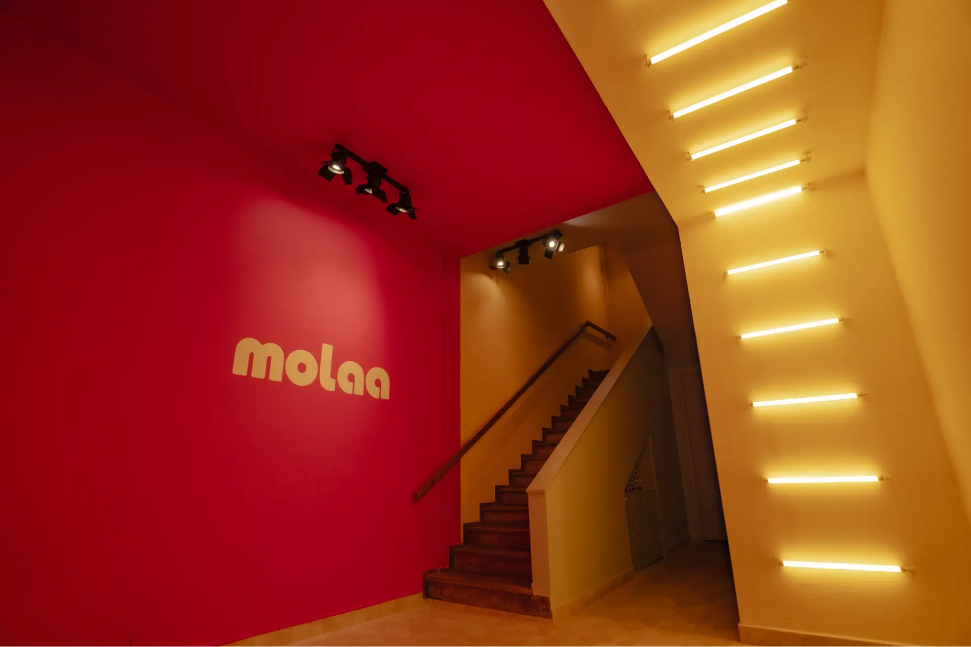 Entrada da sede da MOLAA, com a marca aplicada na parede vermelha e escada iluminada